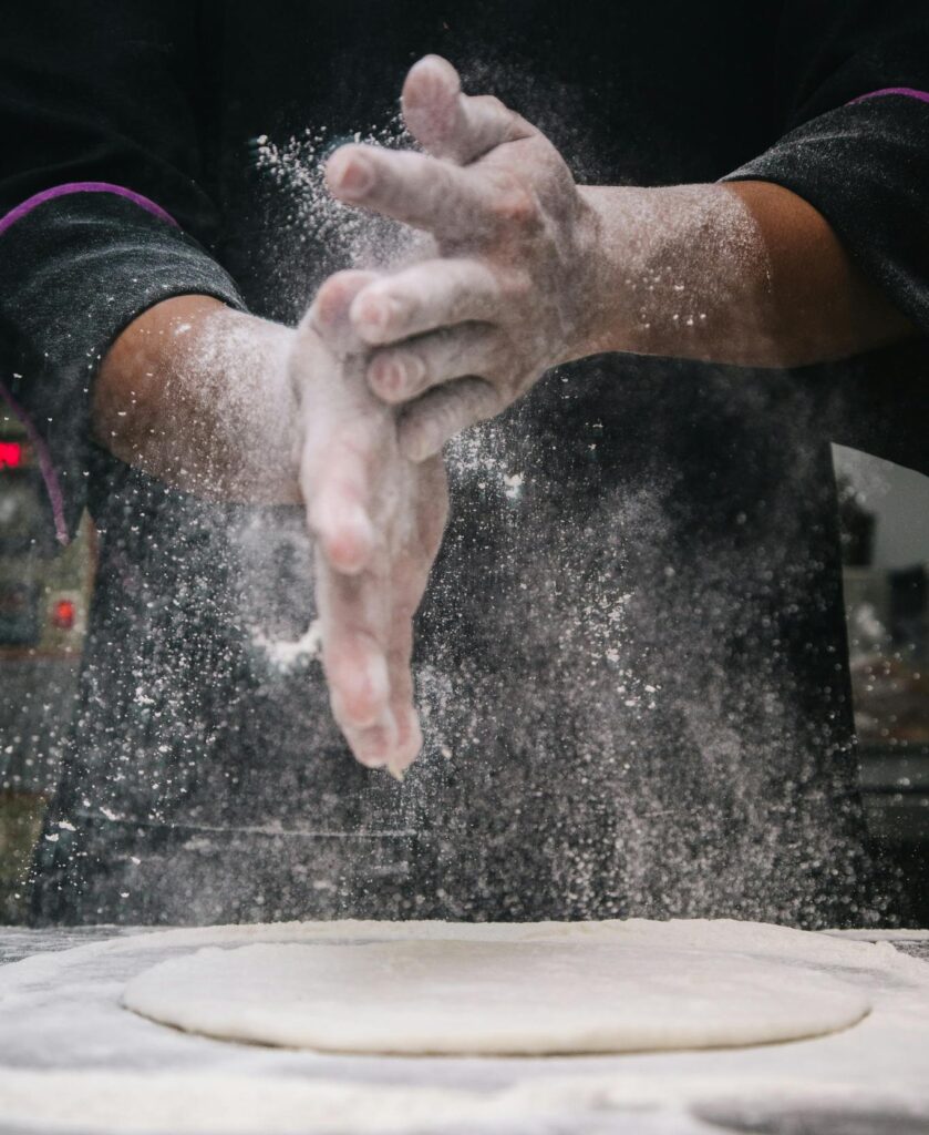 A chef clapping hands over dough, releasing flour in a kitchen setting.